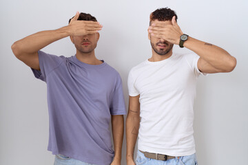 Homosexual gay couple standing over white background covering eyes with hand, looking serious and...