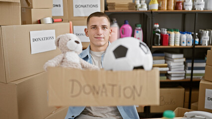 A smiling young man holding a donation box in a storage room filled with various donated items.
