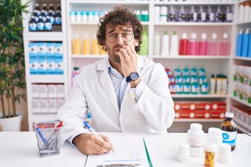 Hispanic young man working at pharmacy drugstore looking stressed and nervous with hands on mouth...