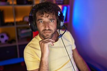 Young hispanic man streamer sitting on table with serious expression at gaming room