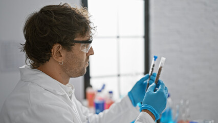 A bearded man in safety glasses and gloves examines test tubes in a bright laboratory setting.