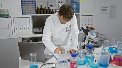 Focused scientist in lab coat analyzing test tubes in a modern laboratory environment, with microscope and laptop present