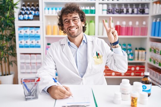 Hispanic young man working at pharmacy drugstore smiling looking to the camera showing fingers doing victory sign. number two.