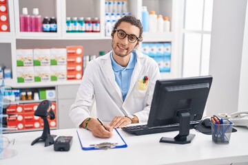 Young hispanic man pharmacist writing on document at pharmacy - obrazy, fototapety, plakaty