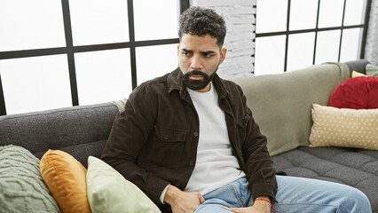 A contemplative man sits on a couch within a well-lit living room, exuding a casual yet stylish...