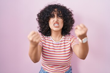 Young middle east woman standing over pink background angry and mad raising fists frustrated and...
