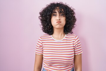 Young middle east woman standing over pink background puffing cheeks with funny face. mouth...