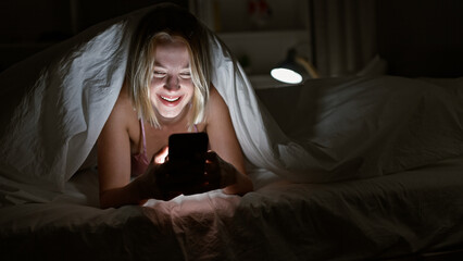 Young blonde woman using smartphone covering with blanket smiling at bedroom - obrazy, fototapety, plakaty