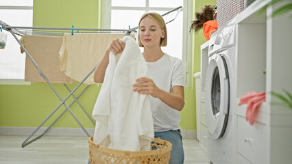 Young blonde woman holding clothes on basket at laundry room