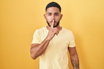 Young hispanic man standing over yellow background asking to be quiet with finger on lips. silence and secret concept.