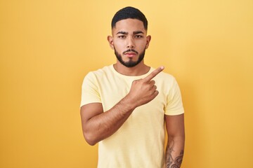 Young hispanic man standing over yellow background pointing with hand finger to the side showing advertisement, serious and calm face