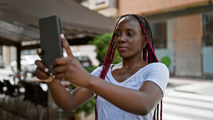 Cool african american woman making a serious selfie with touchpad on a coffee shop terrace in urban...