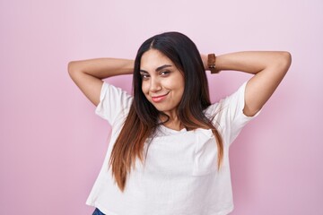 Young arab woman standing over pink background relaxing and stretching, arms and hands behind head...