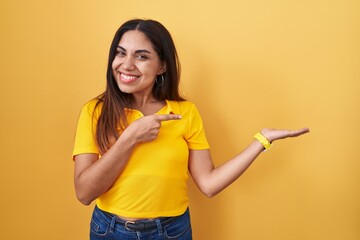 Young arab woman standing over yellow background amazed and smiling to the camera while presenting with hand and pointing with finger.