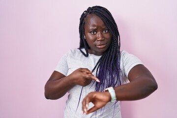 Young african woman standing over pink background in hurry pointing to watch time, impatience,...