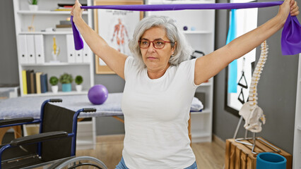 Mature woman exercising with resistance band in rehab clinic alongside wheelchair