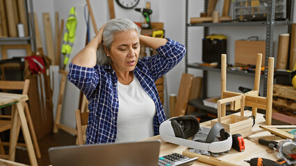 A stressed woman stands in a cluttered workshop, with woodworking tools scattered around her.