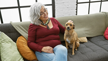Smiling mature woman with glasses pets her brown dog on a couch in a cozy living room.