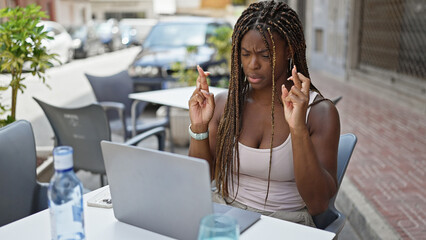 African american woman using laptop crossing fingers for luck at coffee shop terrace