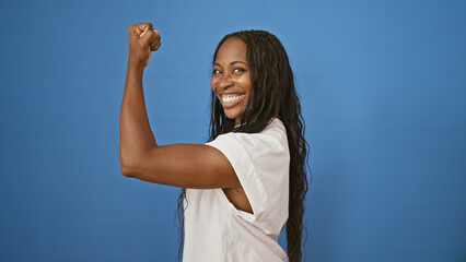 Happy african woman flexing her muscle confidently against a blue isolated background, exuding...