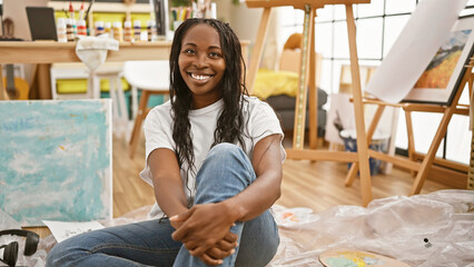 A smiling african american woman artist sitting on the floor in a bright art studio.