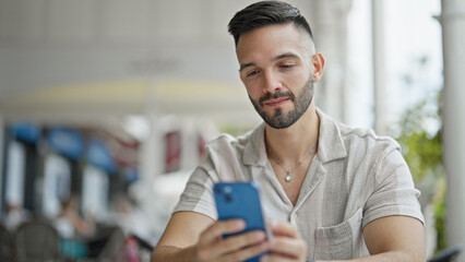 Young hispanic man using smartphone sitting on table at coffee shop terrace
