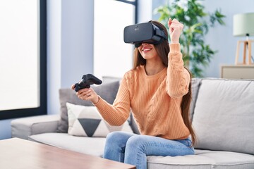 Young beautiful hispanic woman playing video game using virtual reality glasses and joystick at home