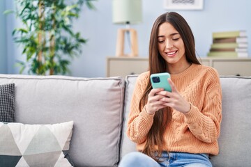 Fototapeta premium Young beautiful hispanic woman using smartphone sitting on sofa at home