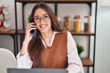 Young beautiful hispanic woman talking on smartphone using laptop at home