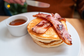 Two crispy bacon strips on a pile of pancakes. There is also a pot of golden maple syrup on the white plate. - 727421595