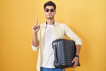 Young hispanic man holding suitcase going on summer vacation pointing up looking sad and upset,...