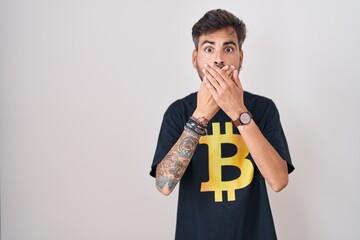 Young hispanic man with tattoos wearing bitcoin t shirt shocked covering mouth with hands for...