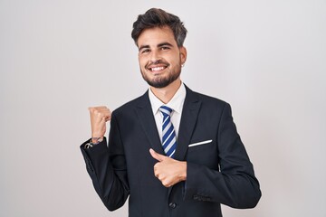 Young hispanic man with tattoos wearing business suit and tie pointing to the back behind with hand...