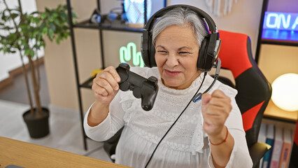 A joyful senior woman with headphones plays video games in a well-lit gaming room, showing...