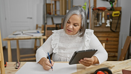 Mature woman using a tablet while writing in a carpentry workshop, surrounded by tools and wood.