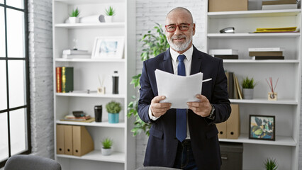Confident middle-aged man with beard and glasses holding papers in modern office.
