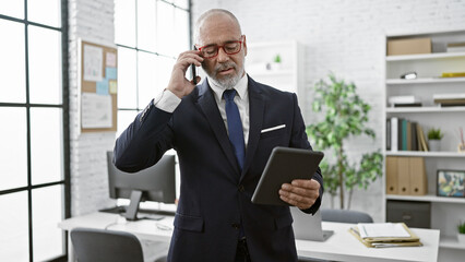 A mature man in a suit uses a tablet and talks on the phone in a bright modern office.