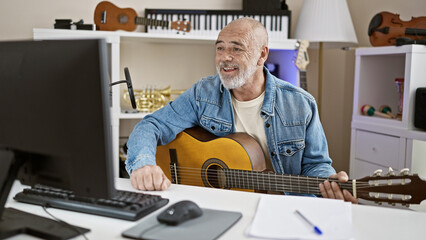 A smiling, mature man in a denim jacket plays guitar in a modern home office with musical...