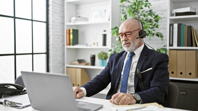 Bald senior man with beard in suit working at laptop in bright office wearing headset