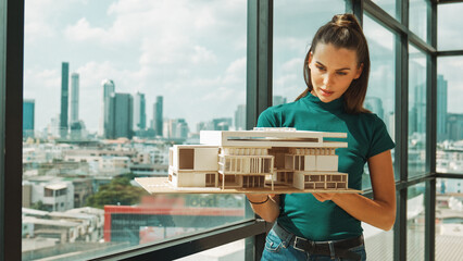 Young smart architect engineer holds architectural model while inspect house model. Professional interior designer checking house construction while standing near window with city view. Tracery
