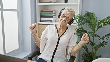 Happy senior woman with grey-hair, confidently dancing to music while working in office, using headphones and laptop