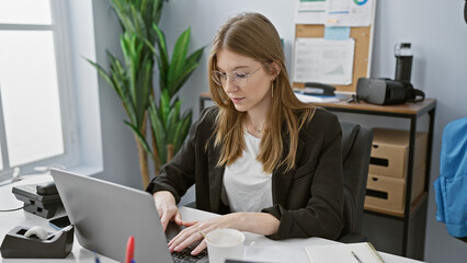 A young adult caucasian woman works diligently in an indoor office setting, looking focused on her...