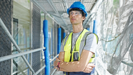 Young hispanic man architect standing with arms crossed gesture and serious face at construction place