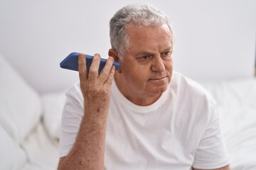 Middle age grey-haired man listening voice message by smartphone sitting on bed at bedroom