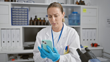 Naklejka premium Beautiful, focused young blonde female scientist immersed in medical research, deftly using her smartphone amidst a slew of test tubes in a bustling, high-tech lab.