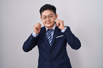 Young asian man wearing business suit and tie covering ears with fingers with annoyed expression for the noise of loud music. deaf concept.