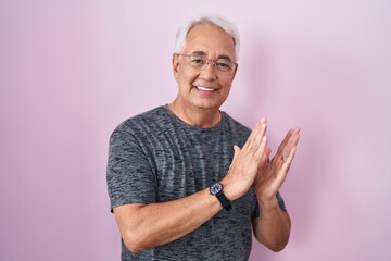 Middle age man with grey hair standing over pink background clapping and applauding happy and...
