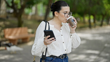 Young beautiful hispanic woman using smartphone drinking take away coffee at the park