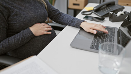 A pregnant hispanic woman working on her laptop in an office, capturing her professional and maternal roles.
