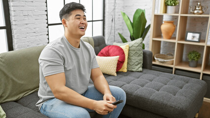 An asian man laughs while holding a remote in a cozy, modern living room.